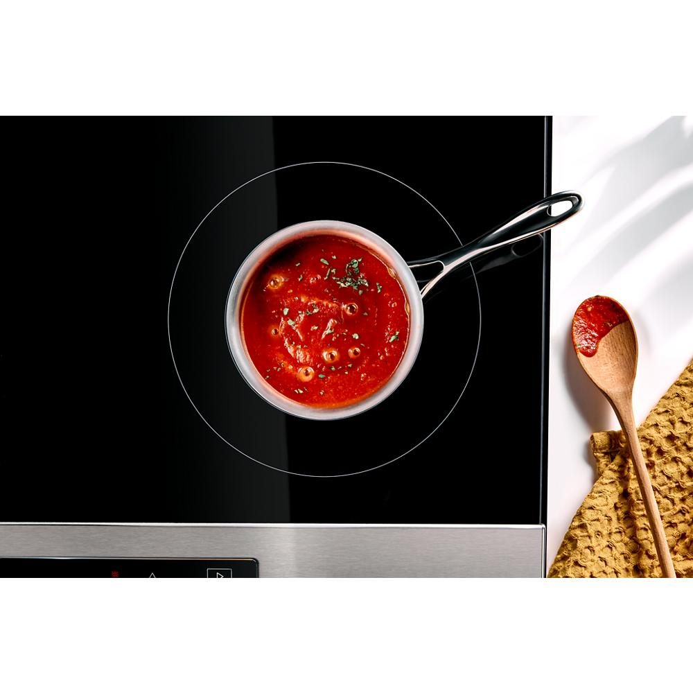 Sauce simmering in a stainless steel saucepan on a black electric induction cooktop with a wooden spoon and kitchen towel nearby