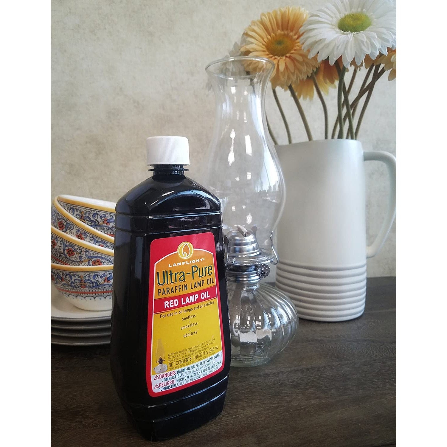 Black plastic bottle of red ultra-pure paraffin lamp oil with white cap beside glass oil lamp and flowers