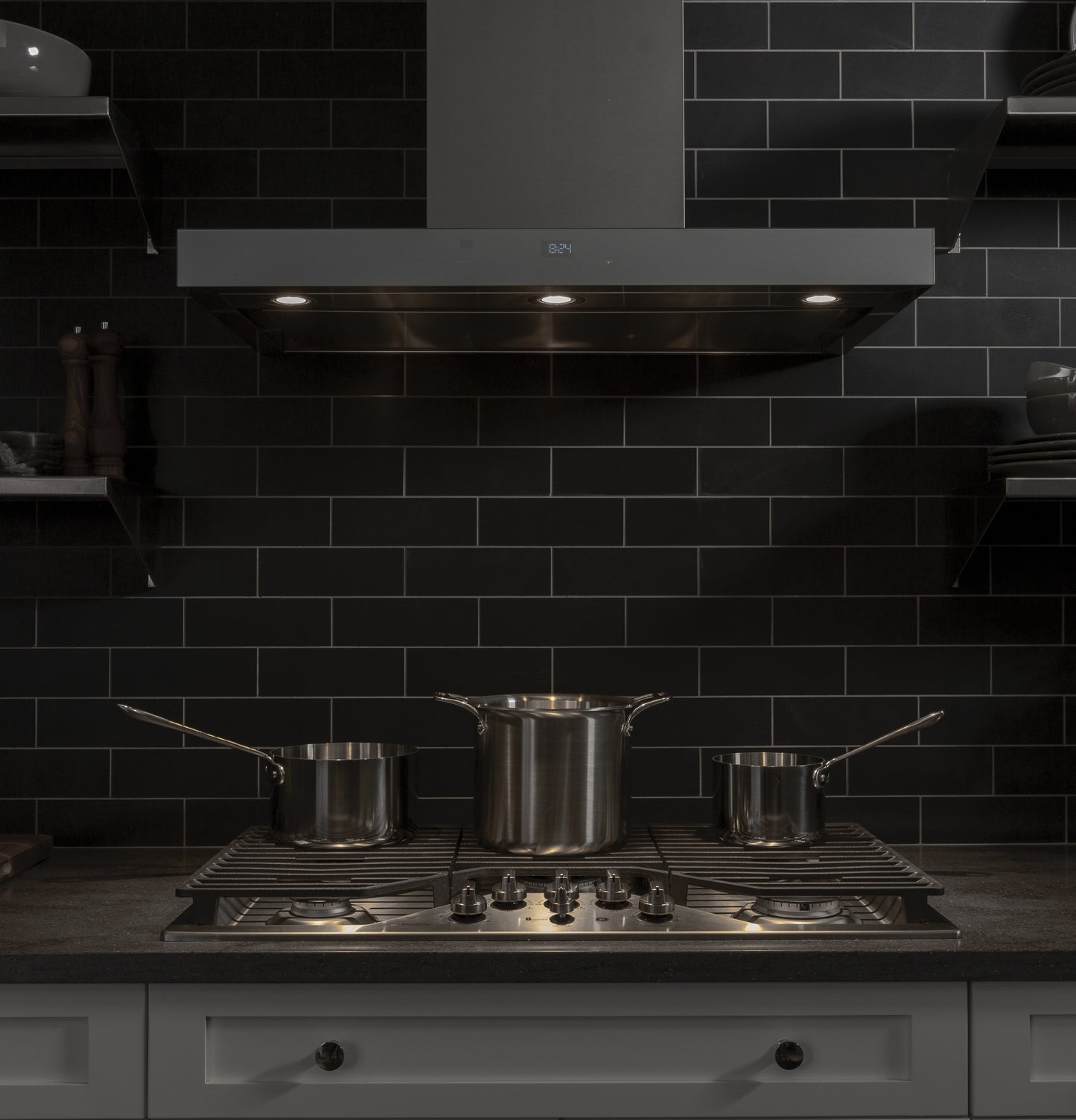 Modern kitchen setup with stainless steel gas cooktop featuring five burners and three saucepans on black tiled backsplash