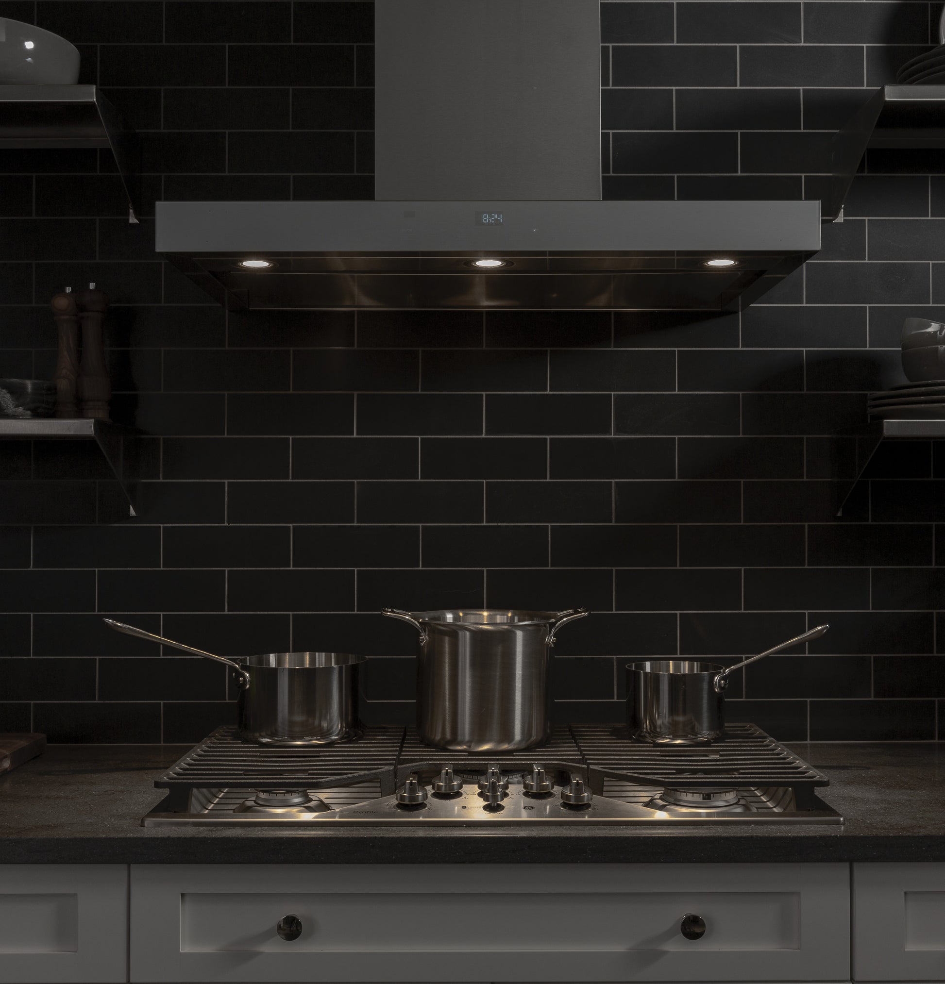 Modern kitchen setup with stainless steel gas cooktop featuring five burners and three saucepans on black tiled backsplash