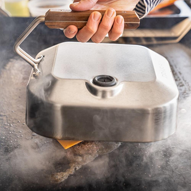 Weighted grill press with wooden handle crushing burgers on griddle for grill accessories and outdoor living use