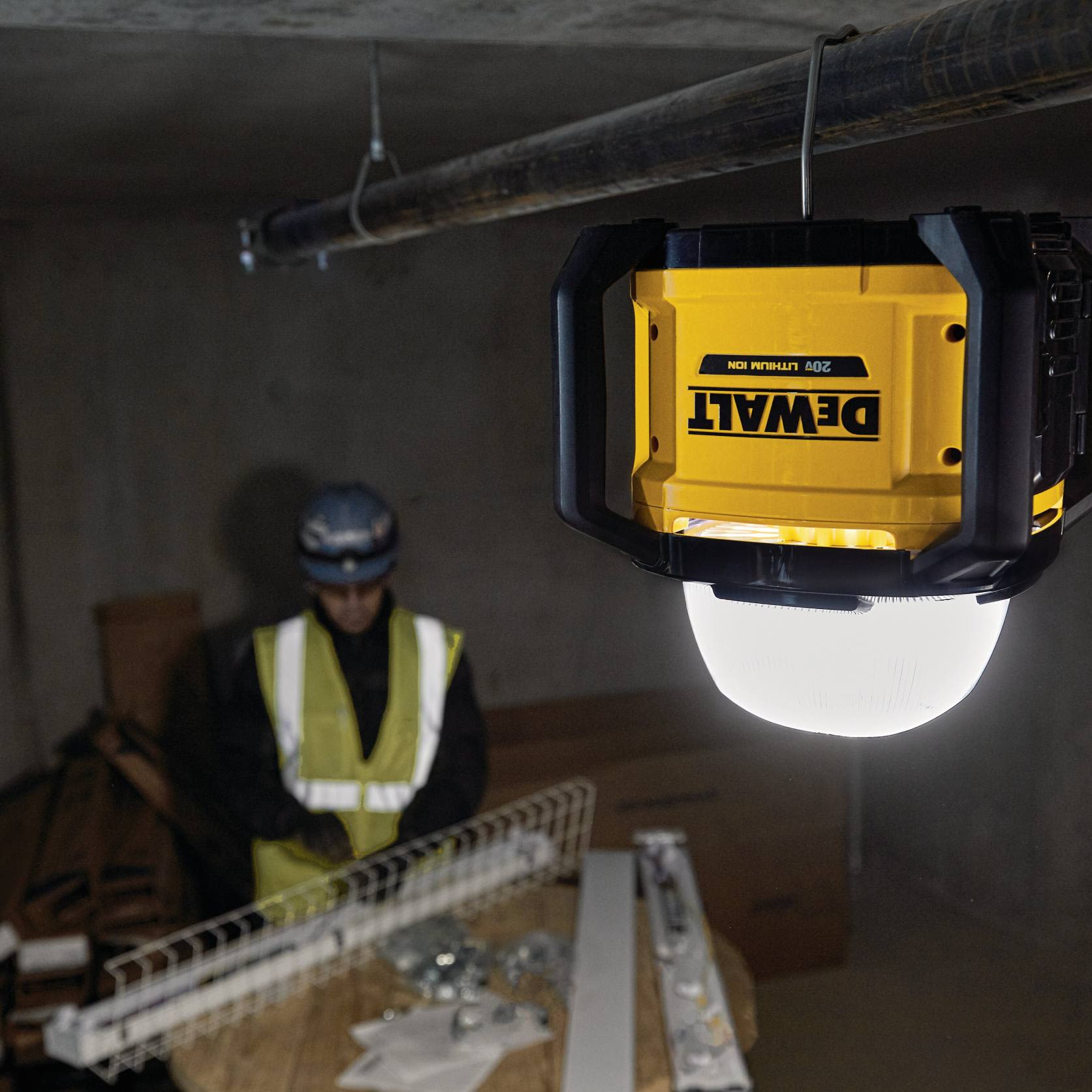 Yellow and black cordless work light hanging from pipe, illuminating construction site with worker in safety vest nearby