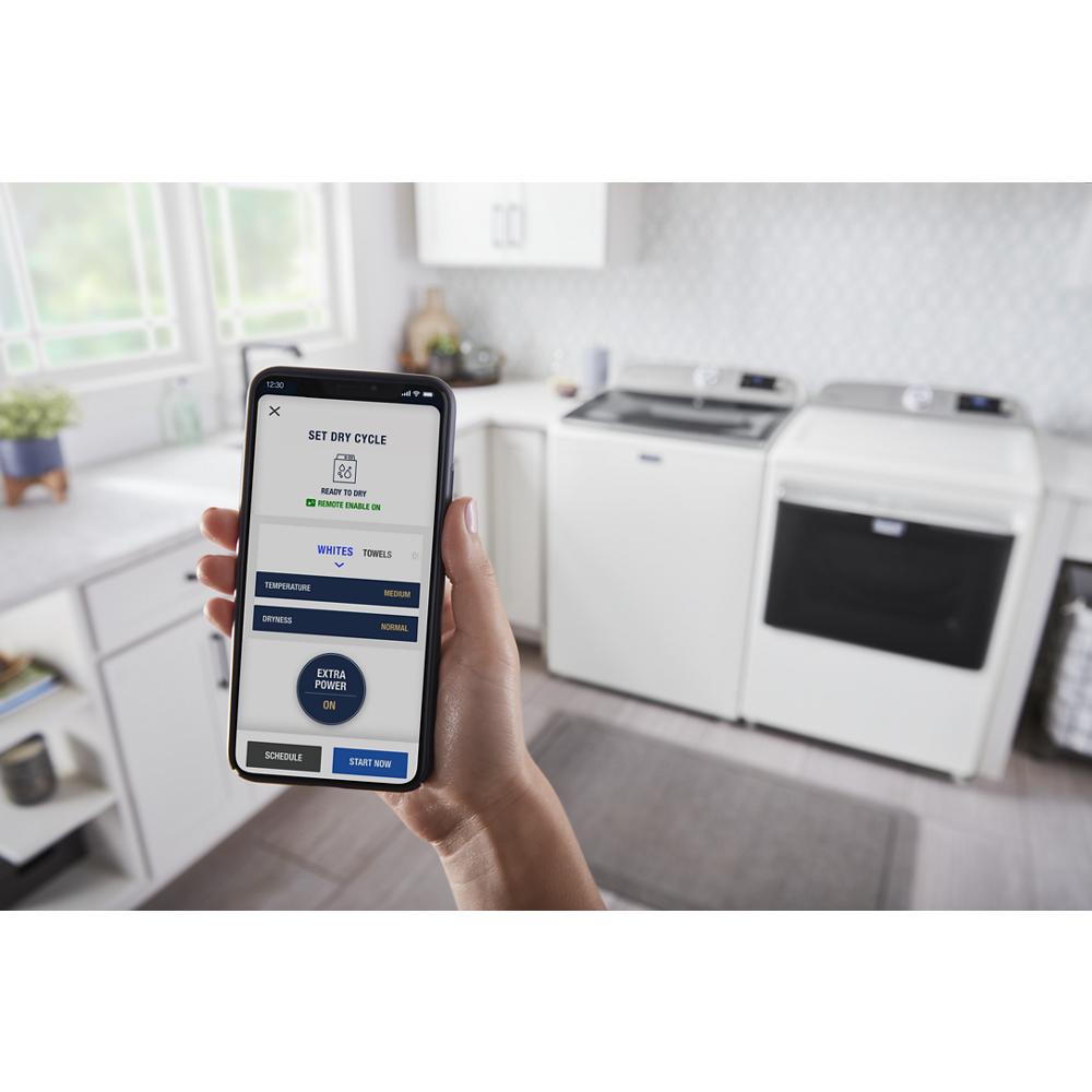 Hand holding smartphone displaying smart dryer app with extra power option in modern laundry room with white appliances