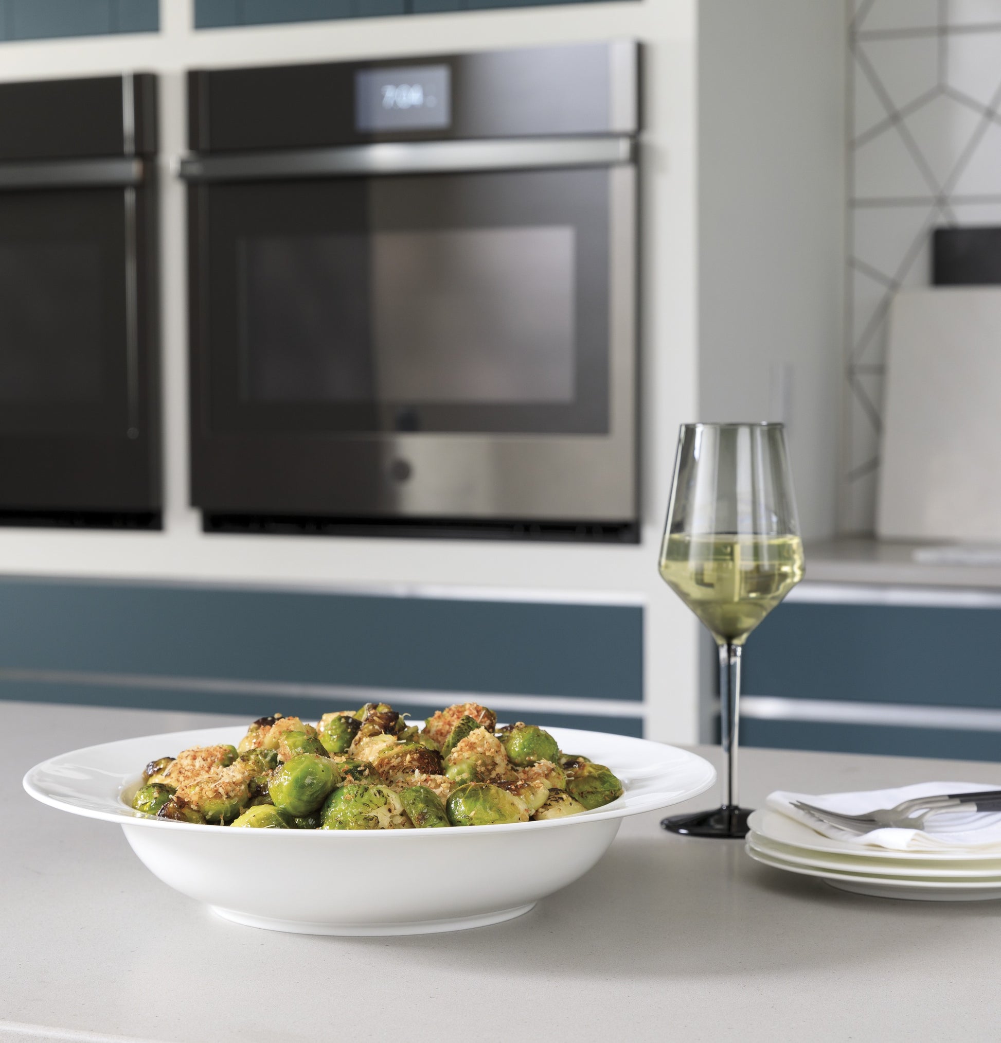 White bowl of roasted Brussels sprouts on kitchen island with glass of white wine and built-in double wall ovens behind