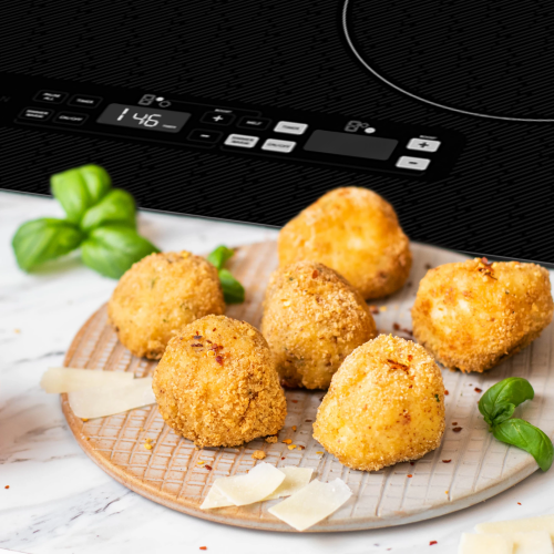 Black induction cooktop with digital touch controls cooking beside plated golden-brown fried croquettes and basil leaves