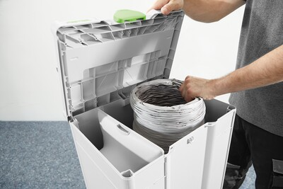 Person loading coiled flexible air supply hose into white portable dust extraction system for woodworking tasks