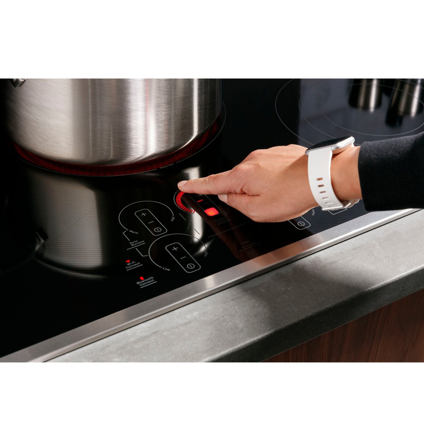 Close-up of hand adjusting touch control panel on black electric built-in cooktop with stainless trim and a pot simmering