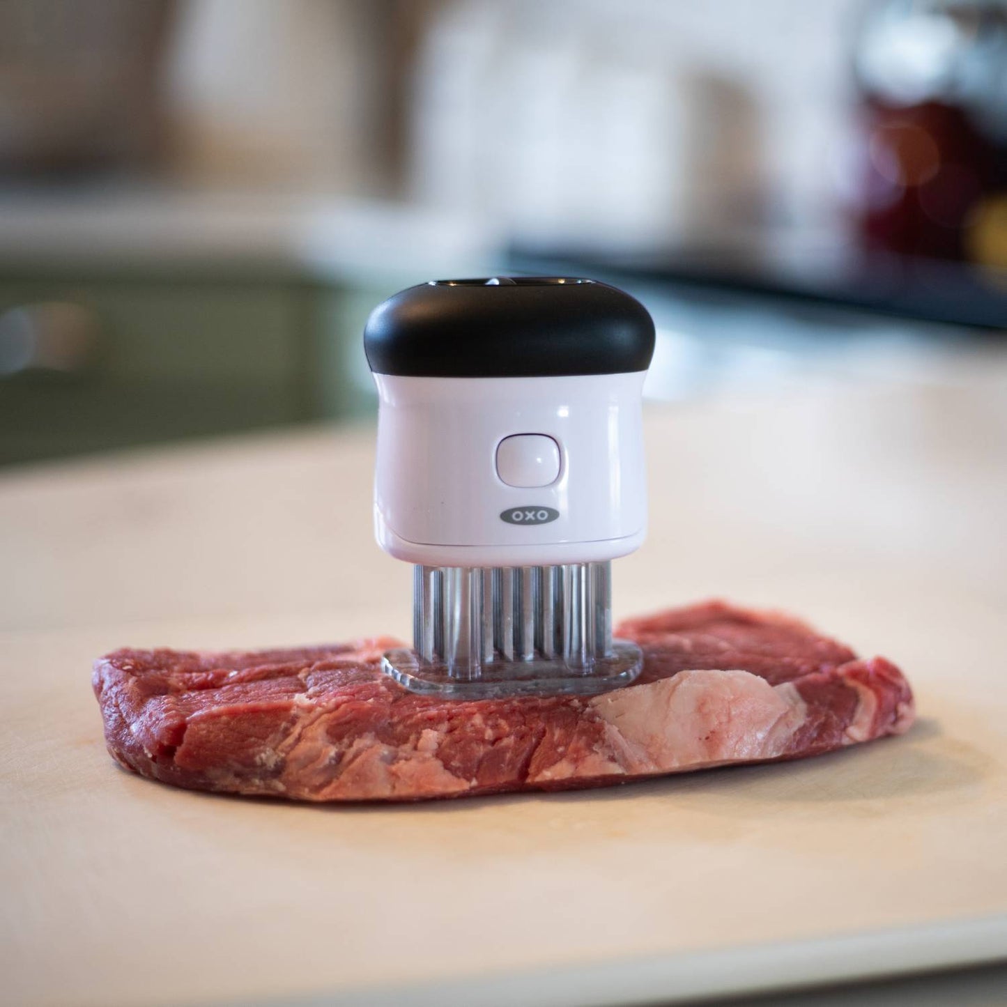 White and black bladed tenderizer pressing into raw steak on countertop in kitchen utensils and cooking tools collection