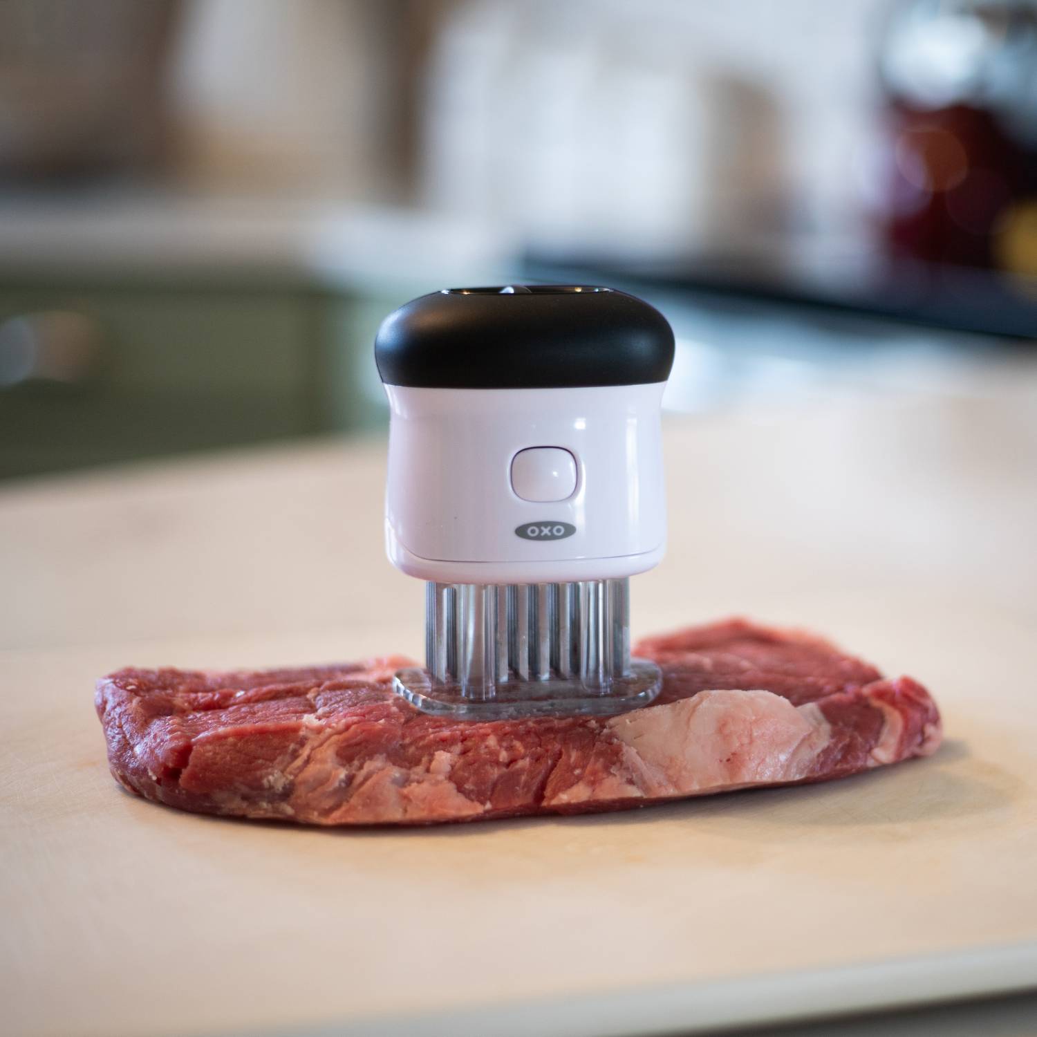 White and black bladed tenderizer pressing into raw steak on countertop in kitchen utensils and cooking tools collection