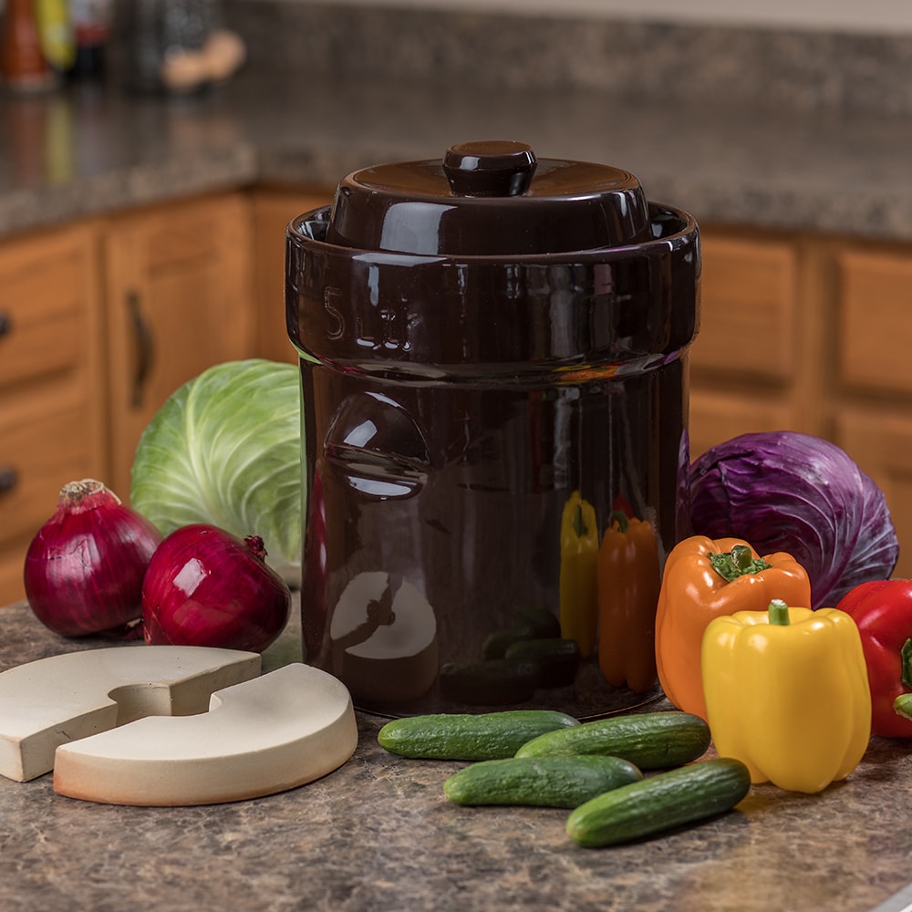 Dark brown ceramic fermenting crock with lid and airlock weights surrounded by fresh vegetables on kitchen counter