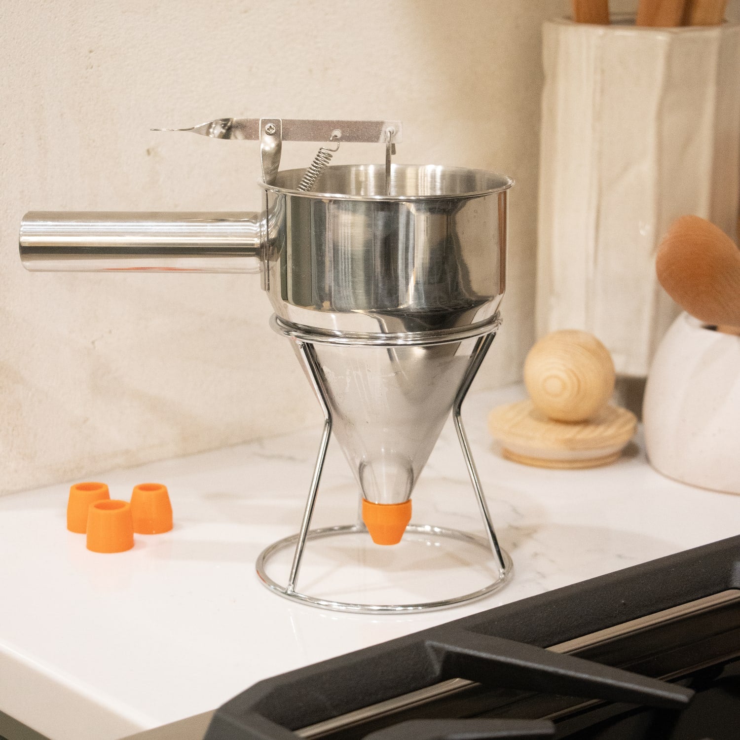 Stainless steel batter dispenser with orange silicone tip resting on metal stand beside extra nozzles on kitchen counter