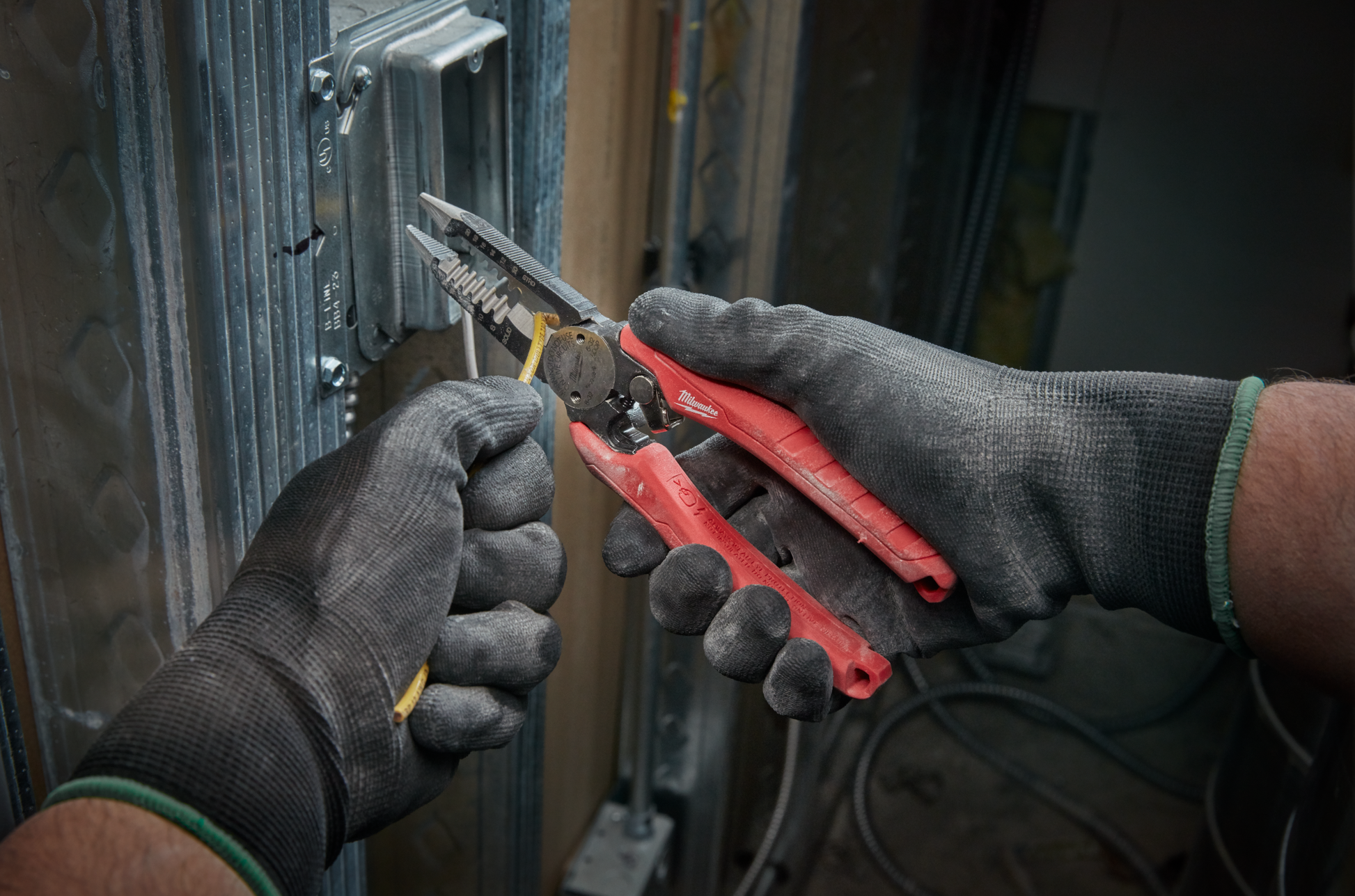 Gloved hands using red comfort grip combination pliers to strip wire inside metal electrical box in home improvement setting