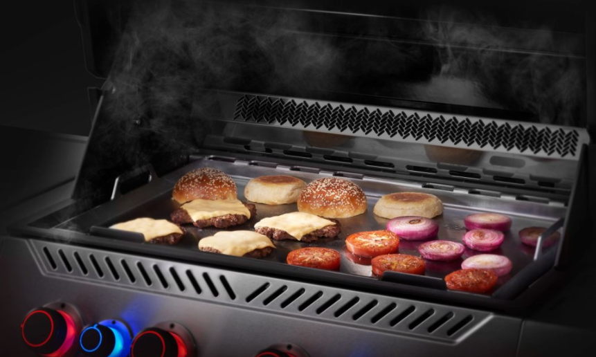 Stainless steel flat top griddle insert cooking burgers, tomatoes, and onions on a gas grill with illuminated control knobs for outdoor living and grilling tools.