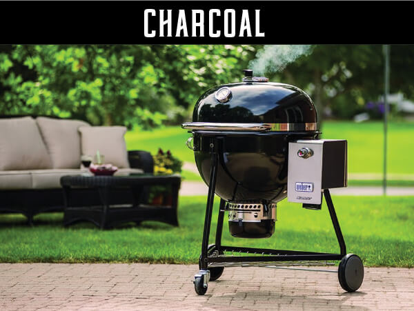 Black charcoal grill with a rounded lid emitting smoke, standing on a patio with green lawn and outdoor furniture in the background.