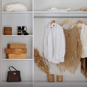 Organized white shelving unit with folded towels, stacked cardboard boxes, black strappy heels, a brown handbag, and a closet rod holding white shirts and a knitted beige shawl.
