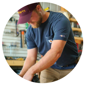 Man wearing a navy blue short-sleeve shirt and maroon cap focused on a woodworking project in a workshop filled with tools and equipment.