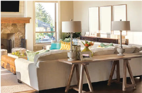 Sunlit living room with a large beige sectional sofa adorned with colorful pillows, a wooden console table with two lamps, and a stone fireplace.