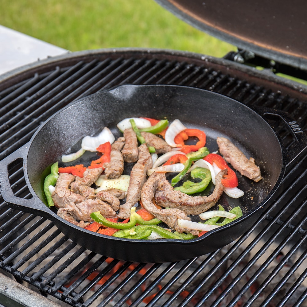 Black cast iron round pan with short side handles cooking seasoned steak strips and bell peppers on grill in kitchenware collection
