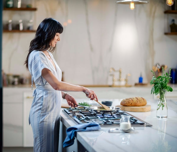Woman stirring food on stainless steel gas cooktop in bright kitchen with fresh herbs and bread nearby