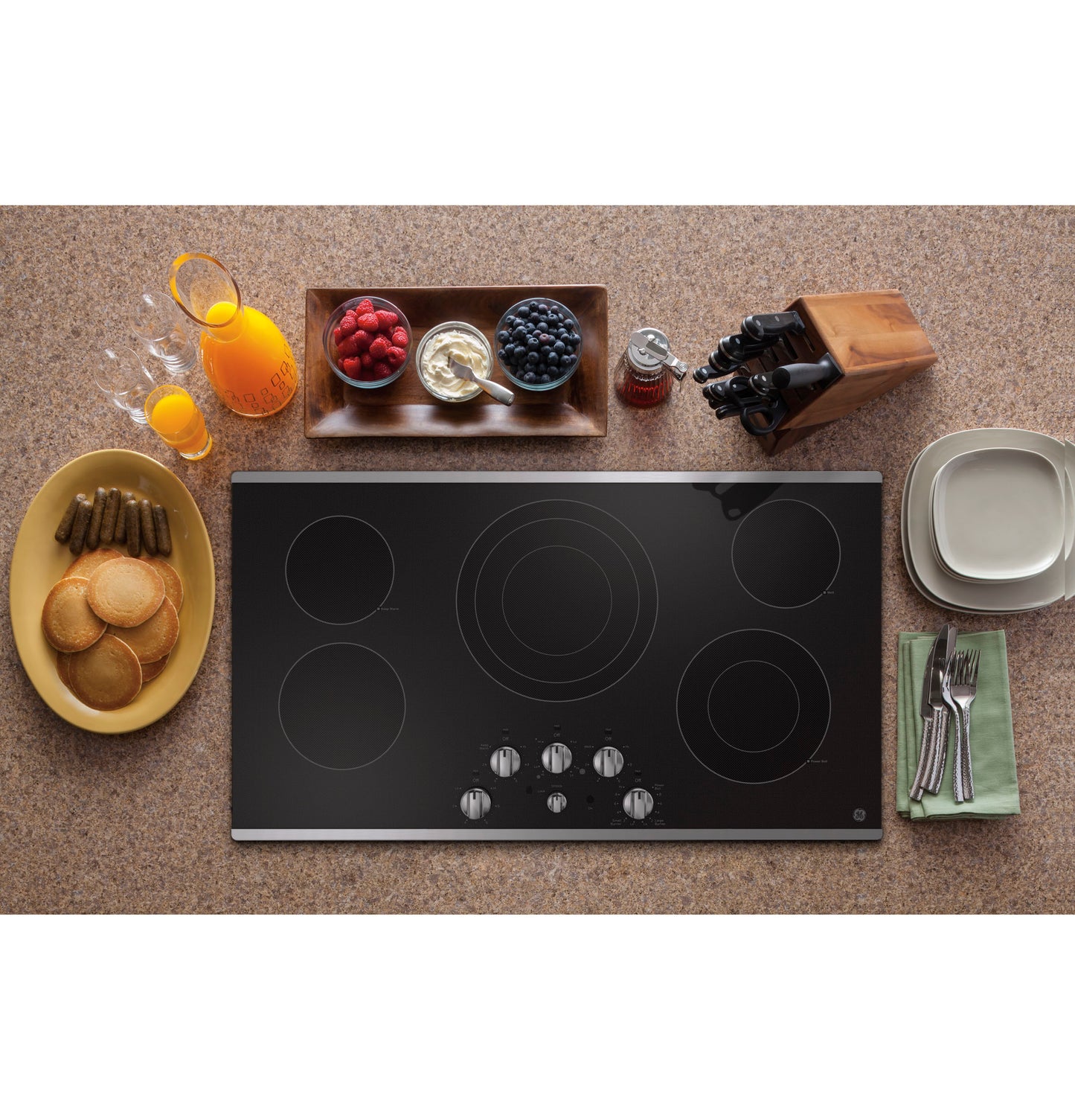 Black electric cooktop with five knob controls surrounded by breakfast setup including pancakes, juice, berries, and cutlery