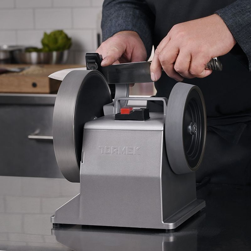 Person sharpening kitchen knife using silver Tormek T-2 Pro sharpener with dual grinding wheels on countertop