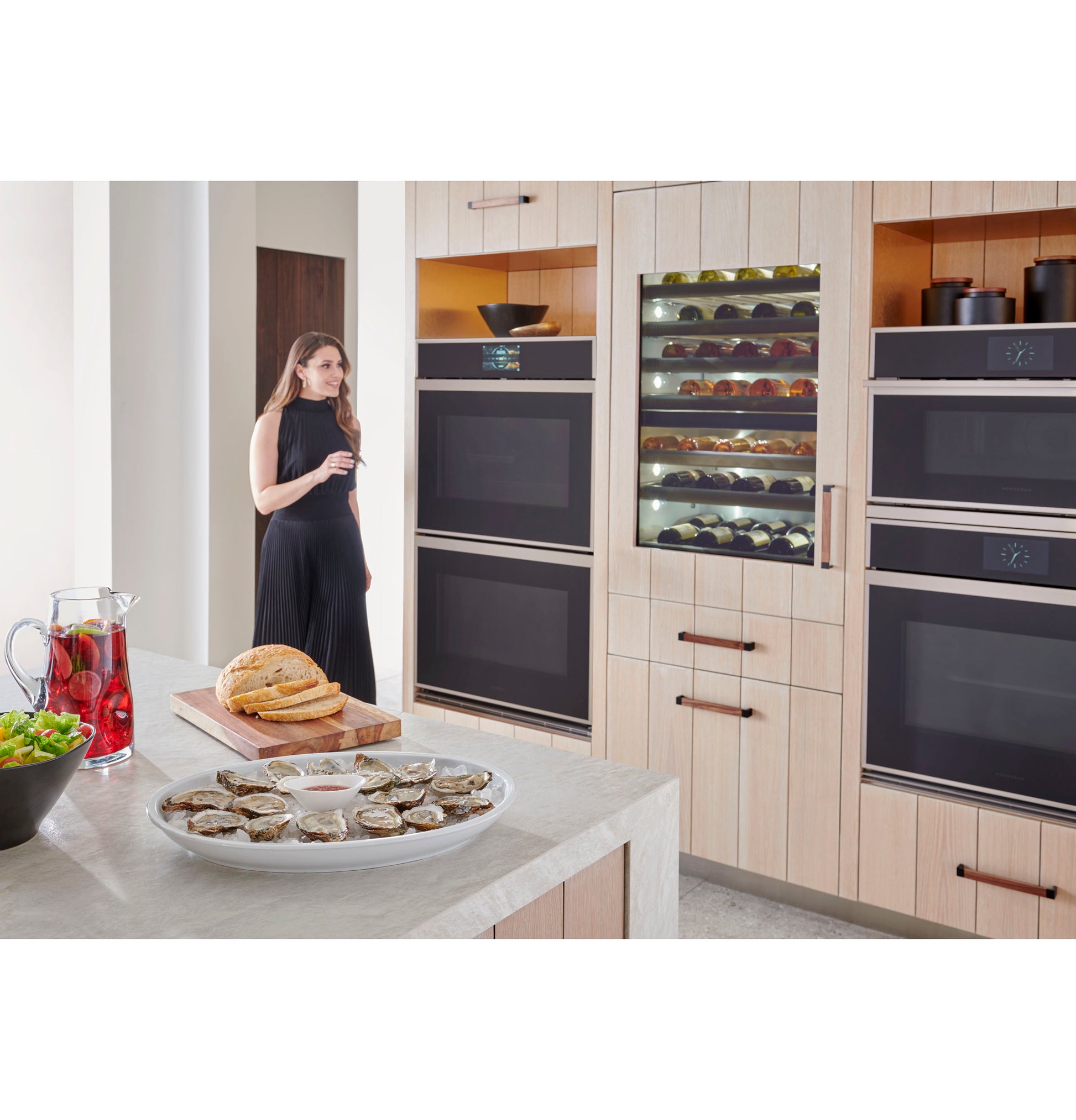 Modern kitchen with Monogram minimalist double wall ovens, built-in wine fridge, light wood cabinetry, and island counter with food
