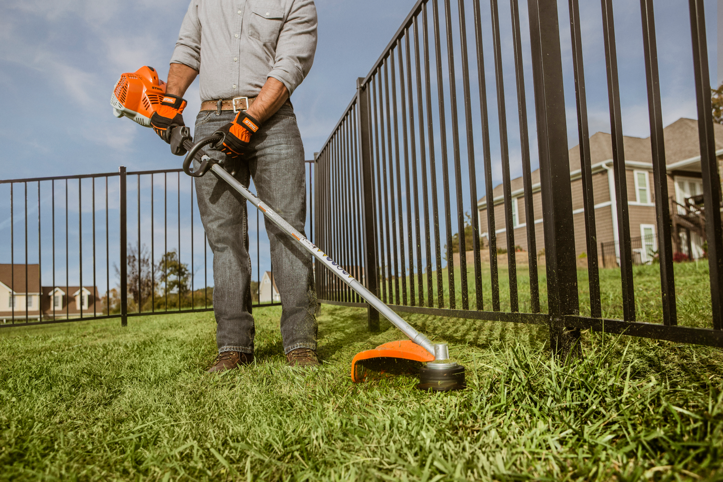 Person using orange and black straight shaft trimmer to cut grass along metal fence in residential yard
