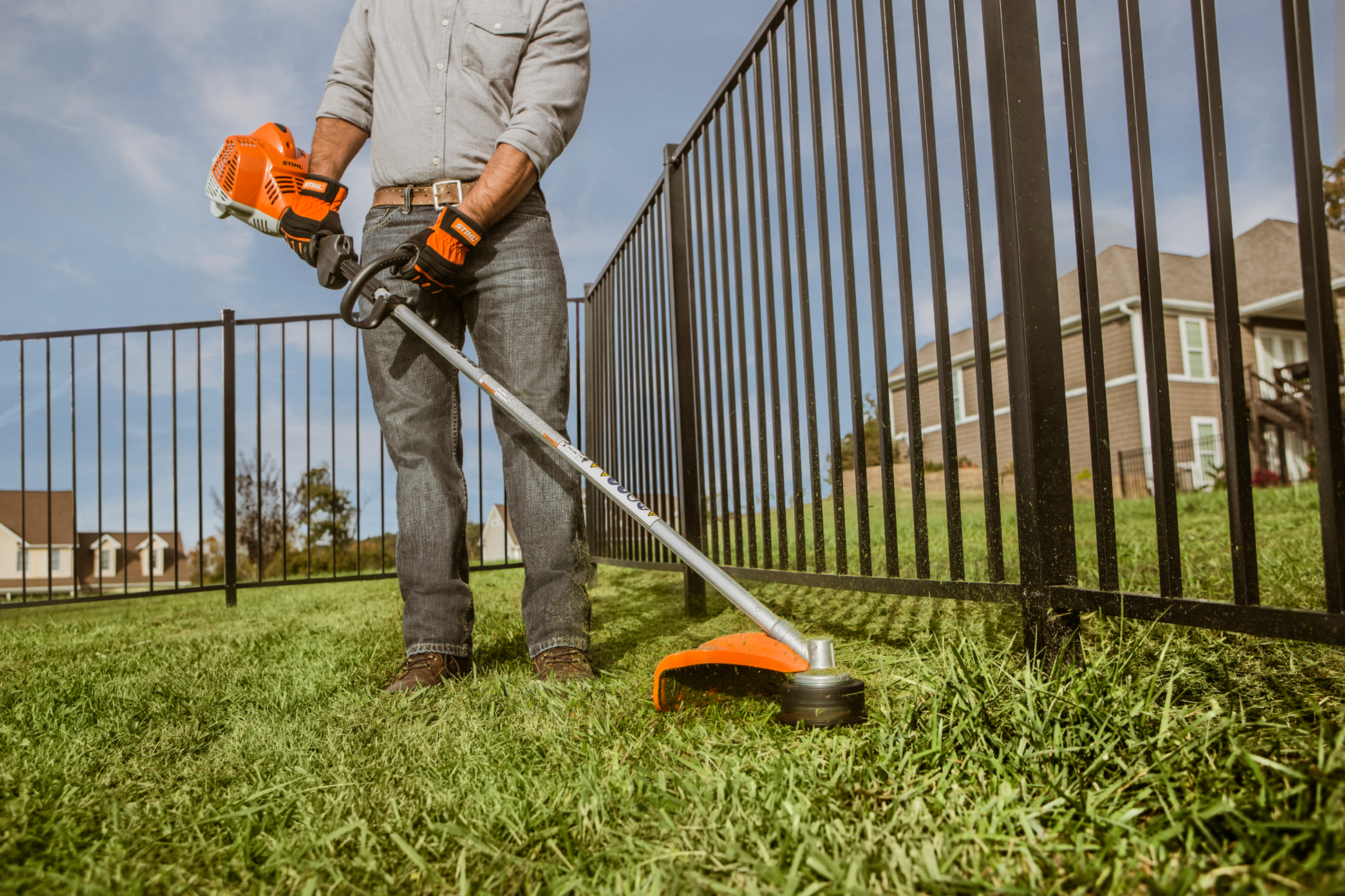 Person using orange and black straight shaft trimmer to cut grass along metal fence in residential yard
