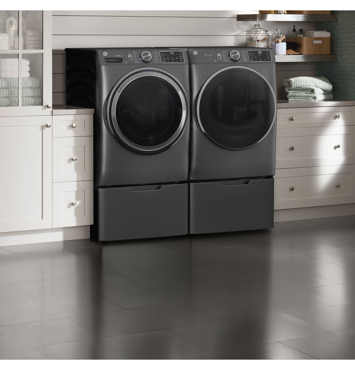 Sleek dark gray front load washer and dryer set with pedestals in a modern laundry room setting with white cabinetry