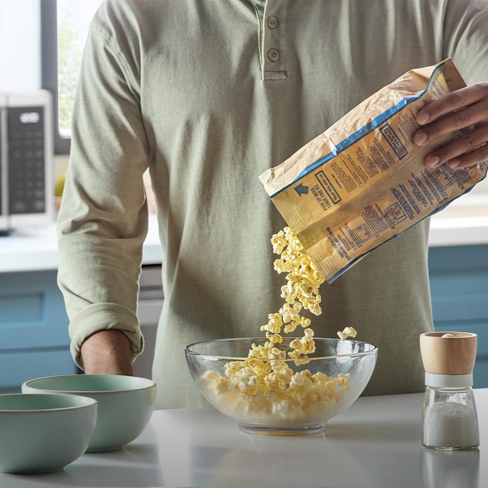 Person pouring freshly popped popcorn into glass bowl on kitchen counter near black countertop microwave