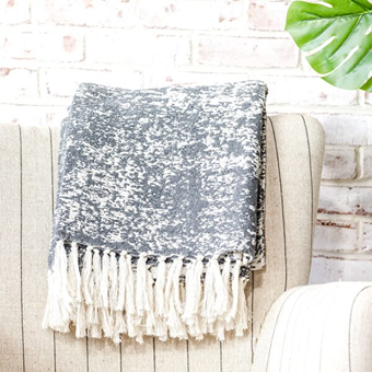 Soft charcoal patterned throw blanket with white fringe draped over a beige upholstered chair in cozy home setting
