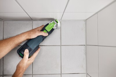 Hands using Festool oscillating multi-tool with round blade removing grout from tiled floor corner for home improvement