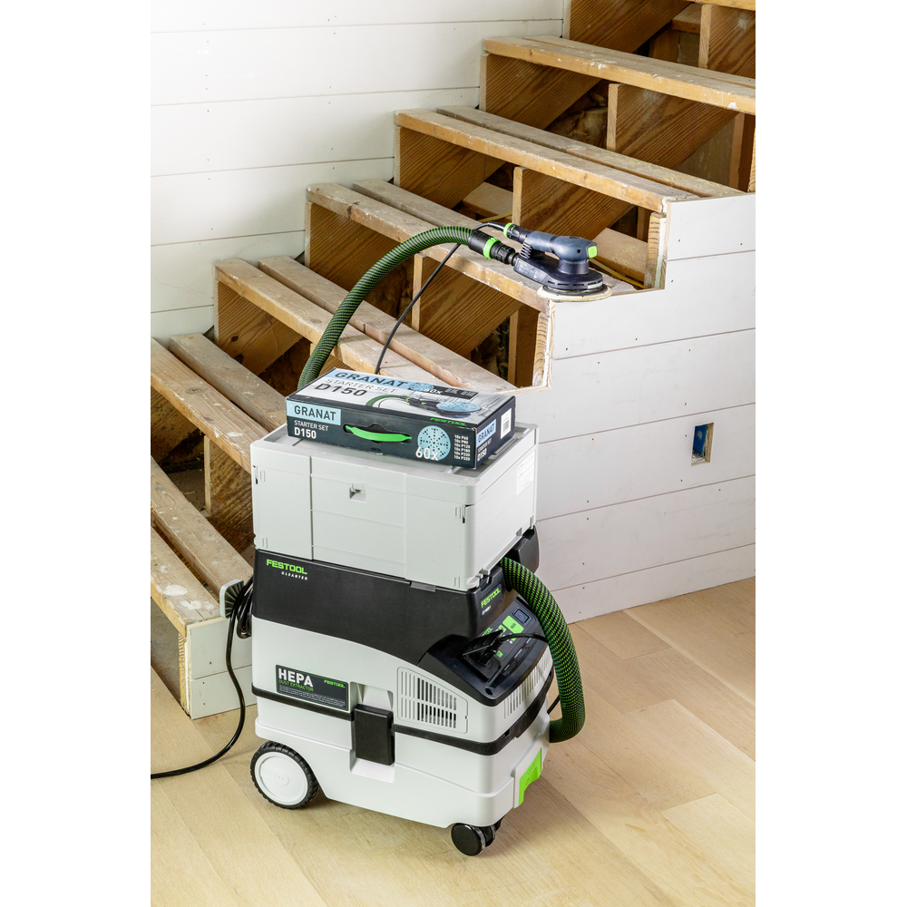 Compact Festool power sander connected to a HEPA dust extractor with sanding supplies on wooden floor by staircase