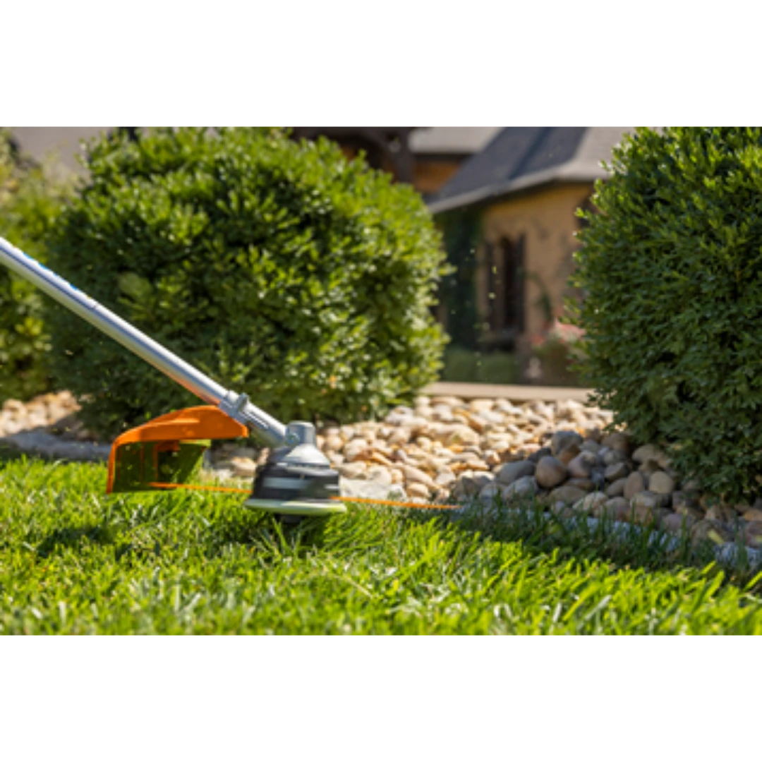 Grass trimmer with silver shaft and orange guard edging green lawn near decorative rock and shrubs for home improvement