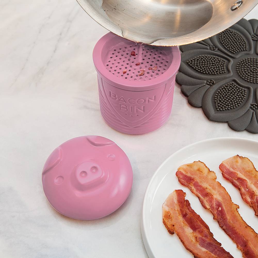 Pink silicone bacon grease holder with pig-shaped lid catching grease from stainless steel pan in kitchenware collection