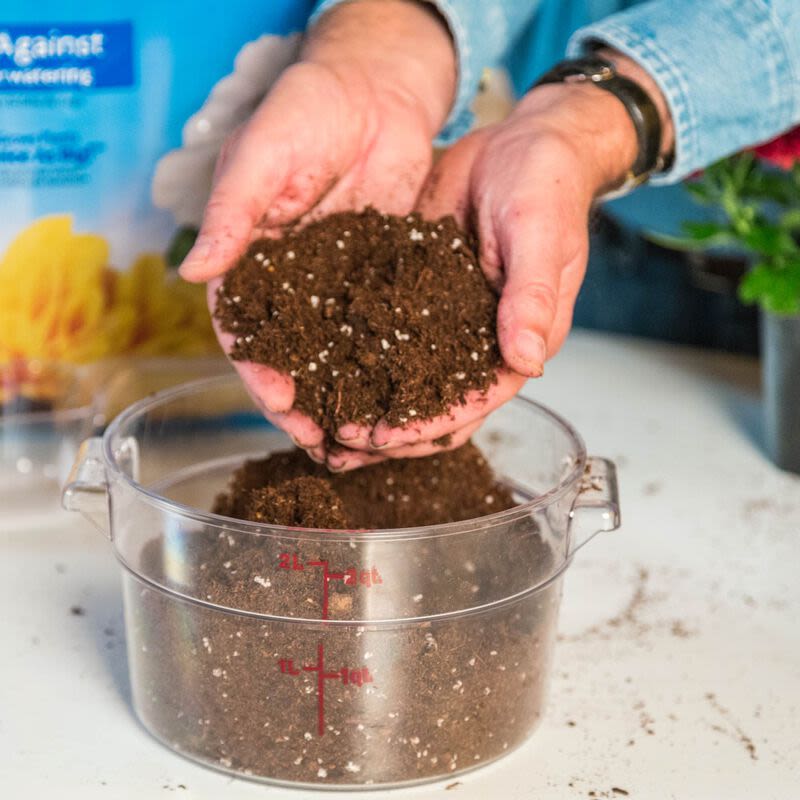 Hands holding dark potting soil with white particles over a clear measuring container on a surface for gardening use