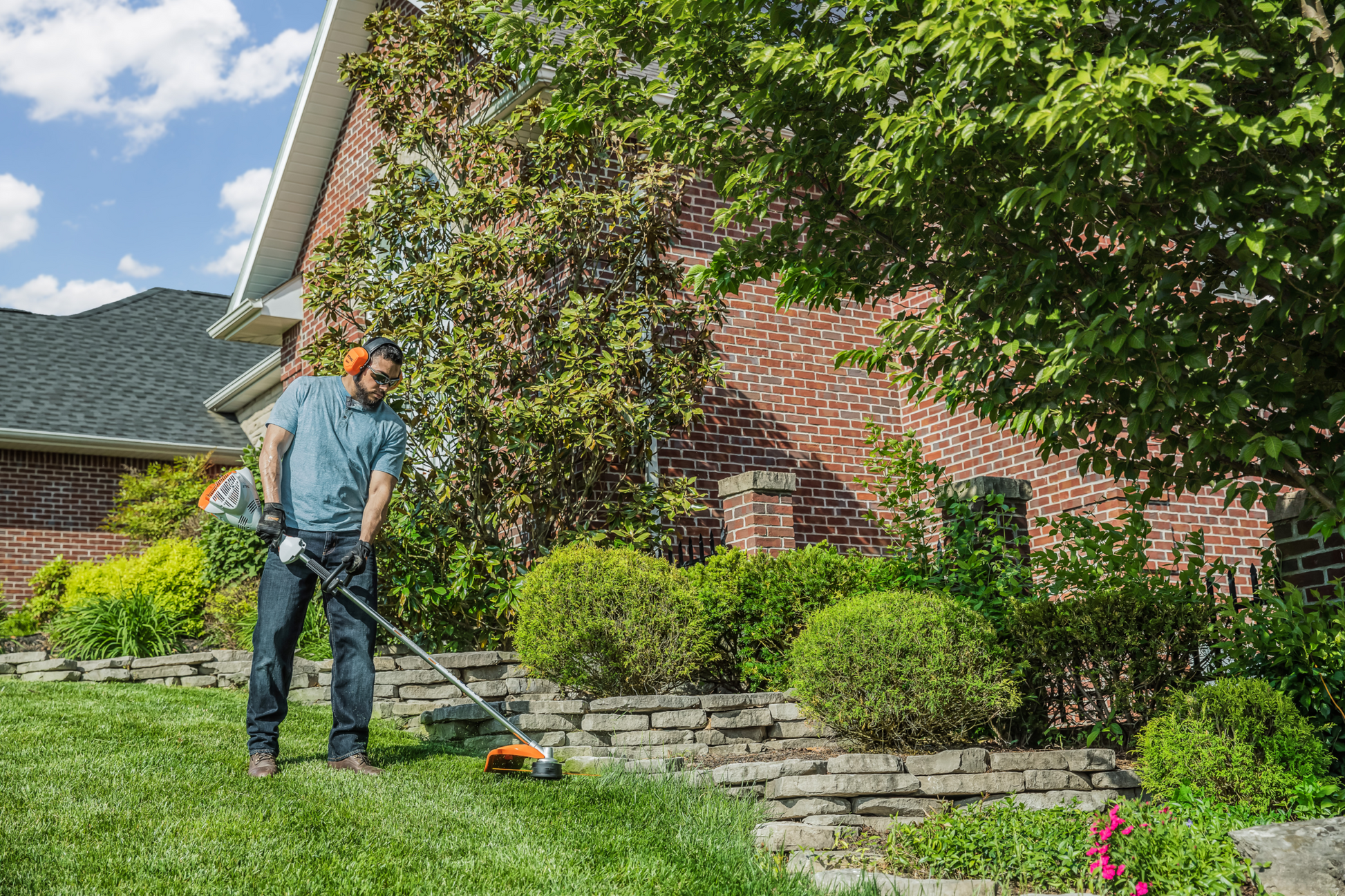 Man using STIHL loop-handle trimmer to maintain a green lawn and garden near a brick house for home improvement