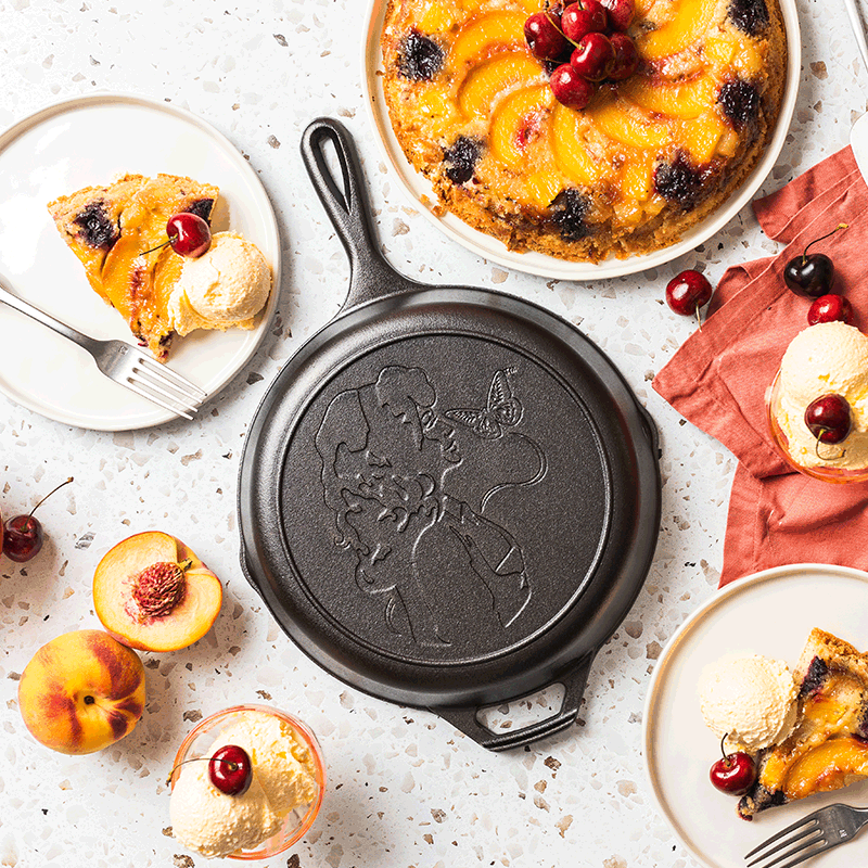 Cast iron skillet with embossed Dolly Parton profile and butterfly, surrounded by peach and cherry dessert, part of Kitchenware collection