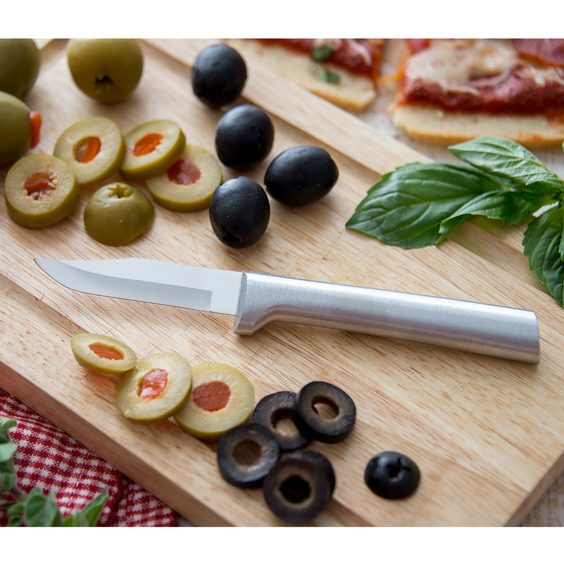 Paring knife with smooth metal handle slicing green olives on wooden board with fresh basil in kitchenware collection