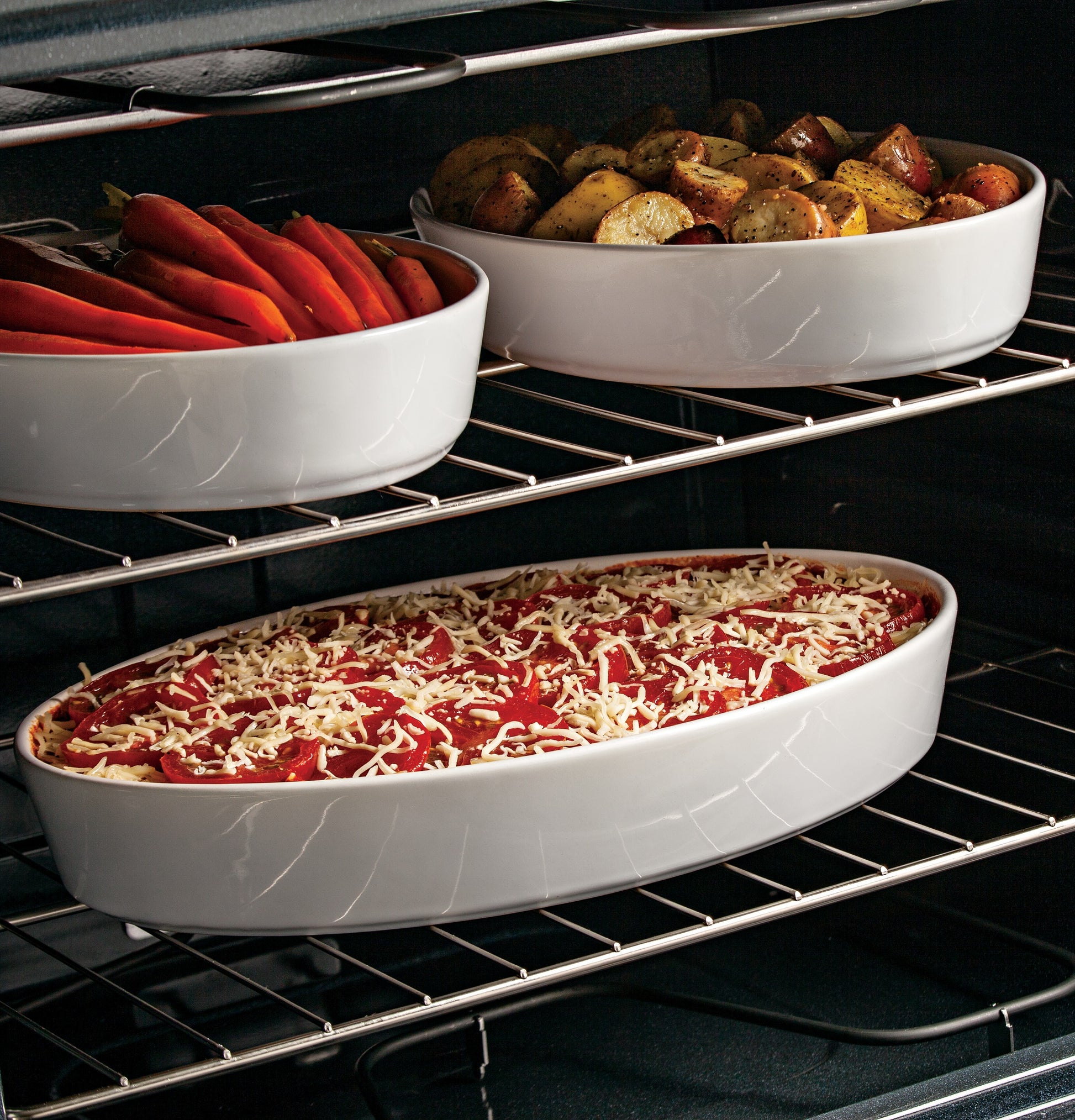 White oval baking dishes with stuffed red peppers, roasted potatoes, and a casserole inside an electric oven range
