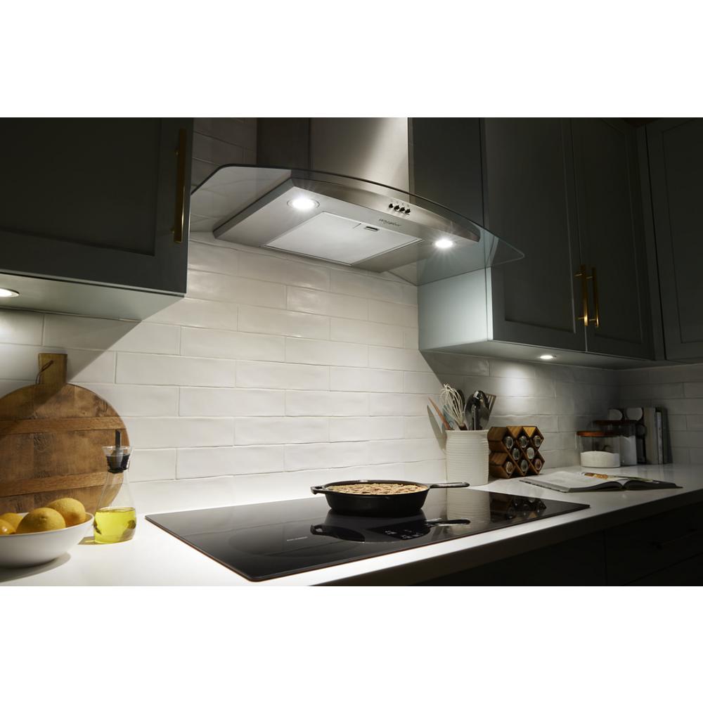 Curved glass wall mount range hood with built-in lights above sleek black cooktop in modern kitchen setting