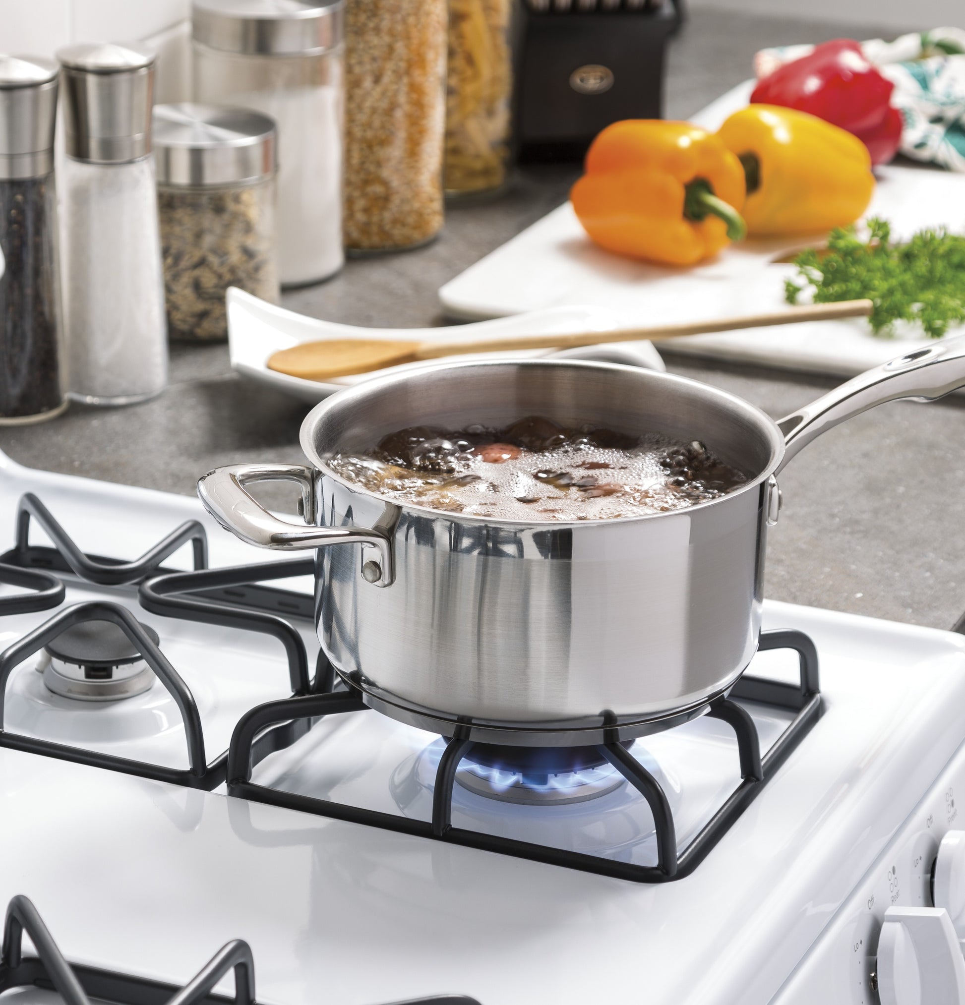 Stainless steel pot boiling on a white freestanding gas range burner with spices and vegetables in kitchen background