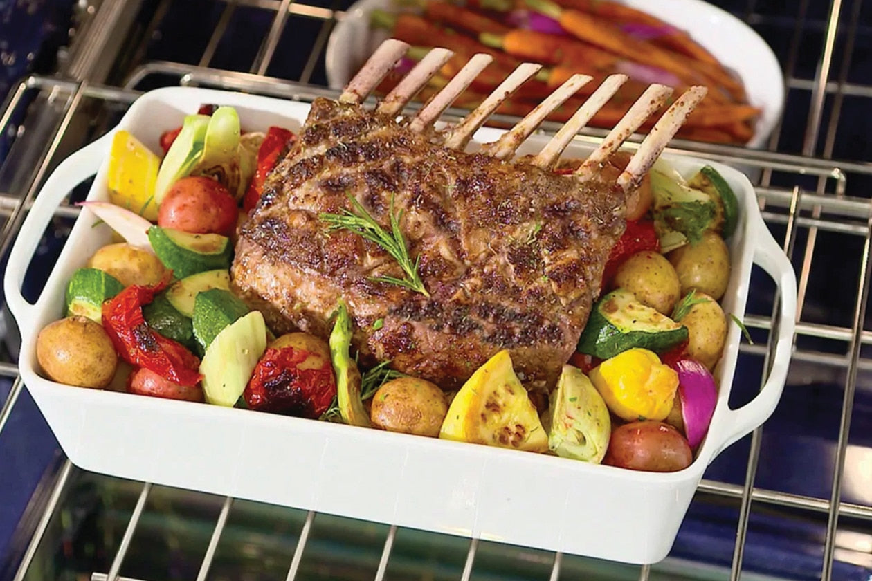 Rack of roasted lamb with assorted grilled vegetables in white ceramic dish inside wall oven