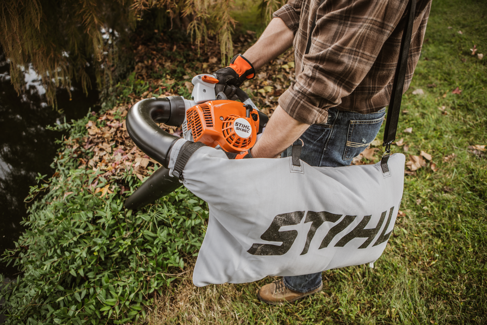 Person using orange and gray STIHL gas blower with collection bag to clear leaves near a garden pond