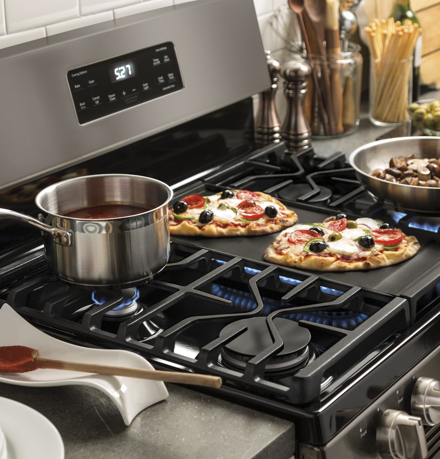 Stainless steel pot simmering sauce and two mini pizzas cooking on a GE gas range cooktop in a kitchen setting