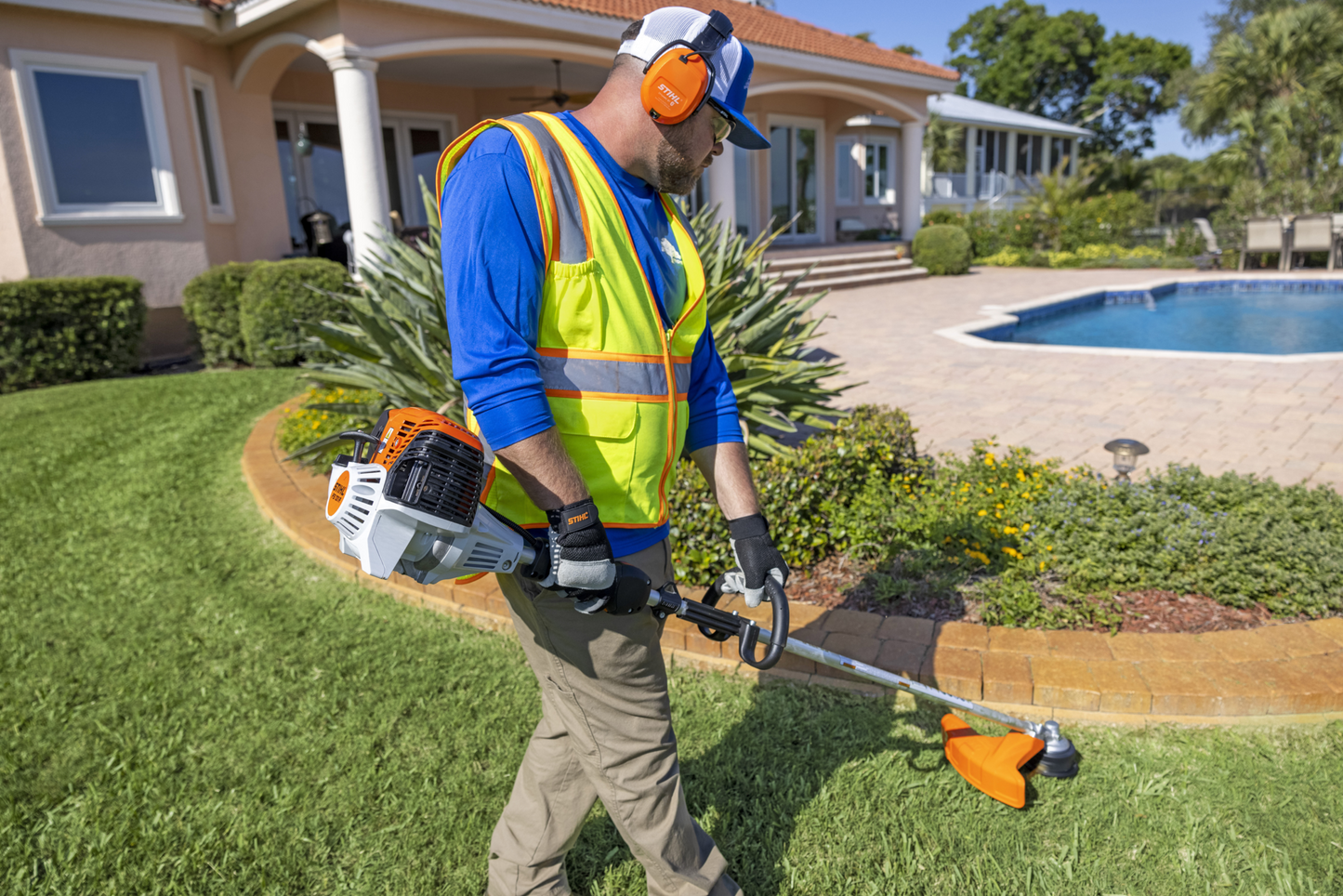 Worker using a professional loop-handle gas trimmer for home lawn maintenance near a residential pool area