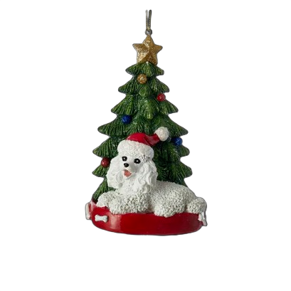 White poodle figurine wearing Santa hat lying in front of decorated green Christmas tree ornament