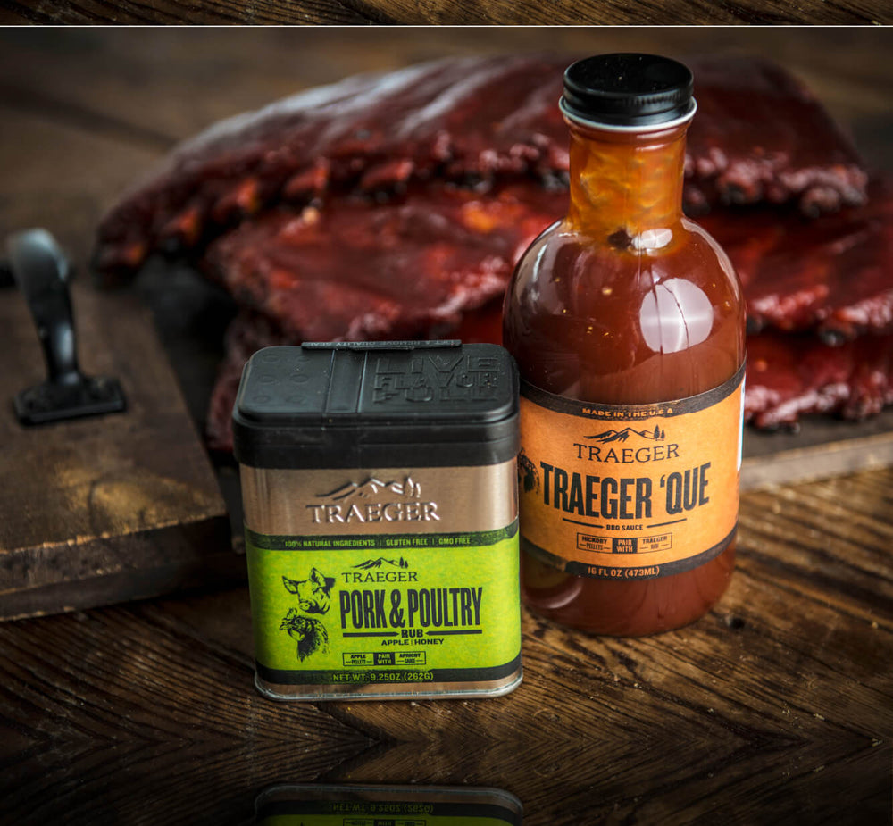 Close-up of a bottle of barbecue sauce and a container of pork and poultry seasoning placed on a wooden surface with smoked ribs in the background.