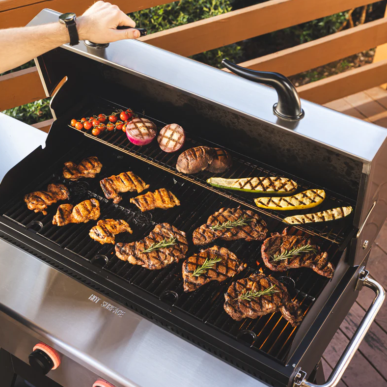 Open pellet grill with assorted grilled steaks garnished with rosemary, vegetables, and bread on dual cooking racks, set outdoors for grilling and outdoor living.