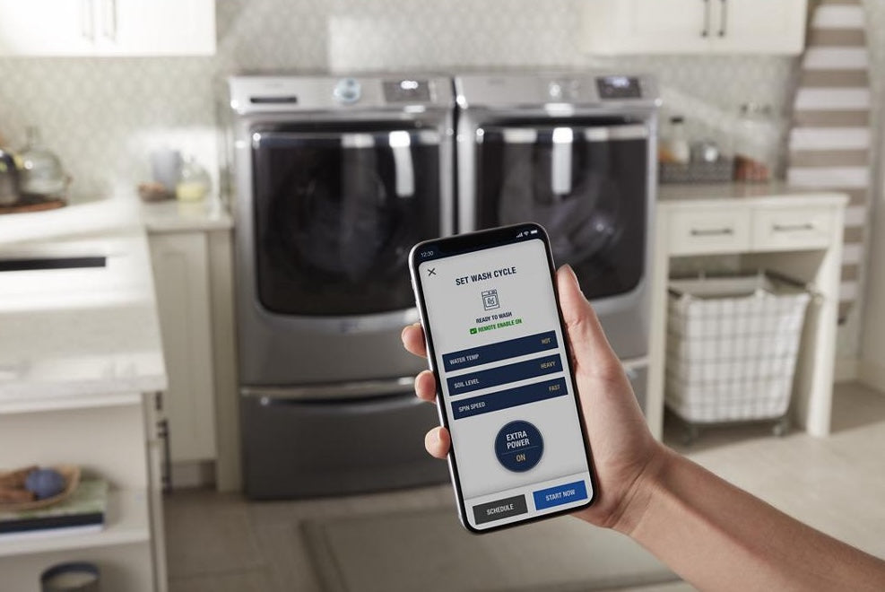 Hand holding smartphone with laundry app in front of matching front load washer and dryer in laundry room